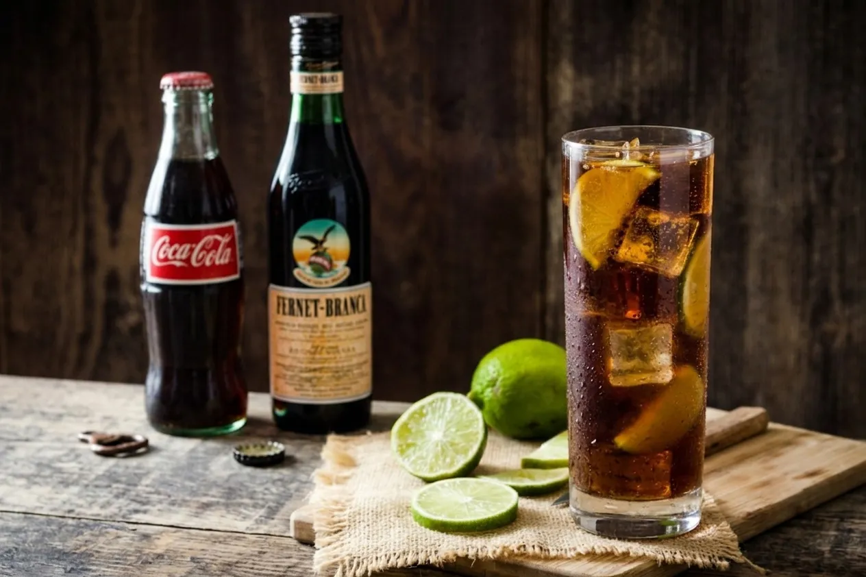 A fernet con coca cocktail served with ice and lime slices.